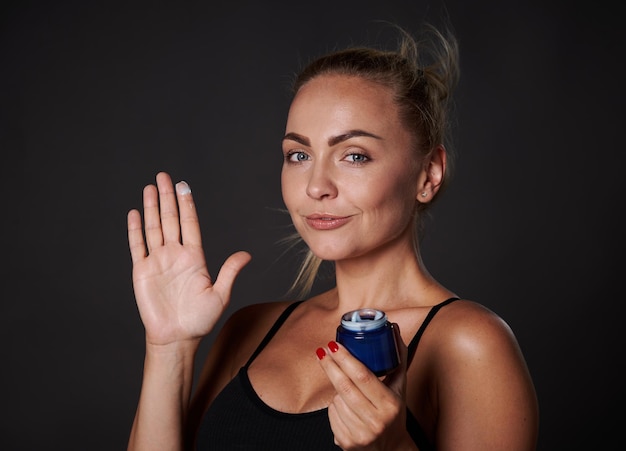 Stunning middle aged Caucasian woman with fresh glowing clear skin smiles looking at camera and holding bottle with antiaging moisturizer facial cream isolated over black background with copy space