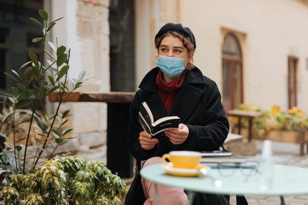 Stunning woman in medical protective mask spending free time at open air terrace and reading new book.