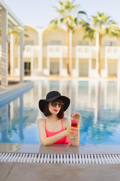 Stunning young lady in trendy pink swimsuit sitting on the edge of pool and enjoying cocktail