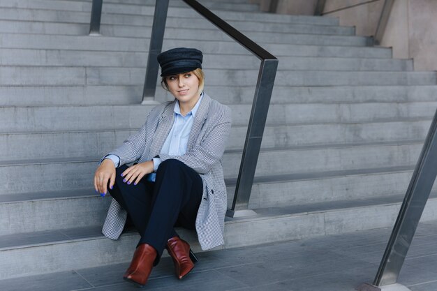 Stunning young lady wearing stylish clothing and cap sitting on stairs near modern building. Concept of urban lifestyle and fashion outfit.