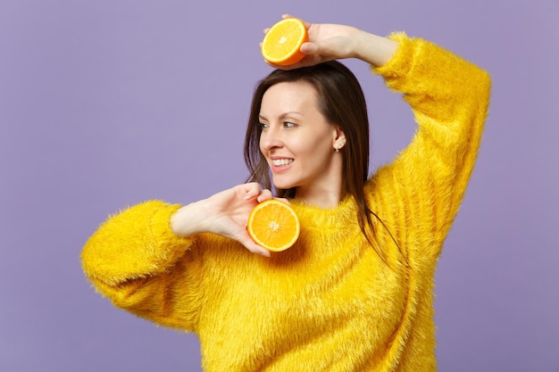 Stunning young woman in fur sweater looking aside holding halfs of fresh ripe orange fruit isolated on violet pastel wall background. People vivid lifestyle relax vacation concept. Mock up copy space.