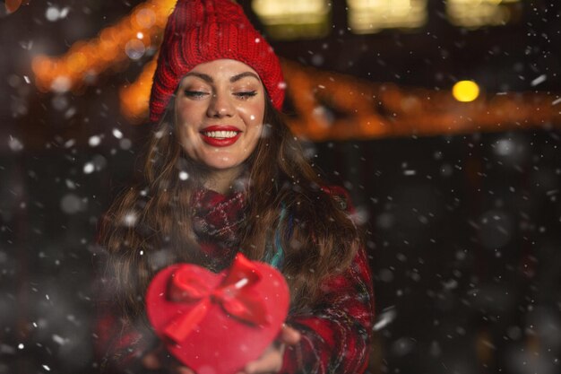 Stunning young woman holding red gift box in a heart shape at the evening during snowfall