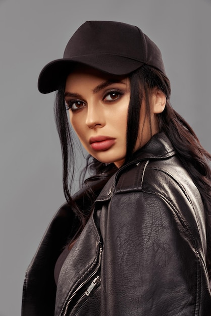 Stunning young woman with make up and trendy hairstyle looking at camera with confidence in eyes. Fashion model wearing black cap and leather jacket. Isolated over grey studio background.