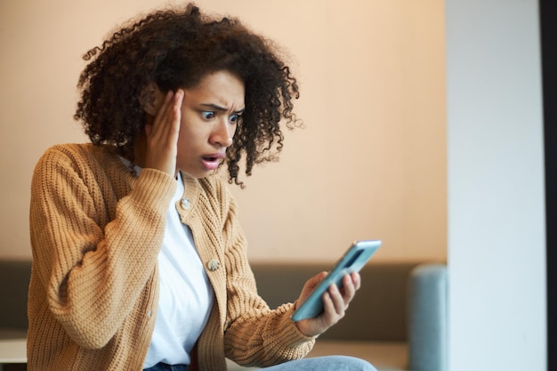 그녀의 휴대 전화를 확인하는 동안 충격과 경악을 표현하는 어리둥절한 아프리카 계 미국인 여성