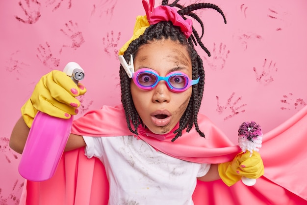 Stupefied female superhero with dreadlocks engaged in household chores holds cleaning detergent and dirty brush busy doing washing has funny look isolated over pink wall