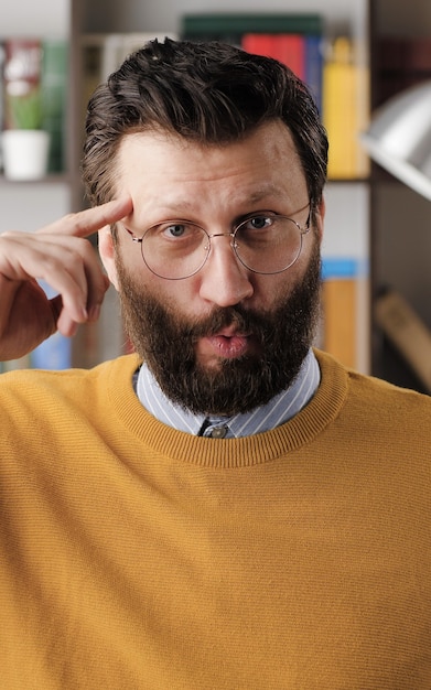 Тупой. Раздраженный бородатый мужчина в очках в офисе или квартире смотрит в камеру и крутит пальцем у виска. Вид крупным планом