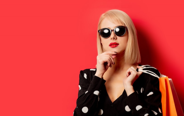 Style blonde girl in sunglasses with shopping bags on red wall