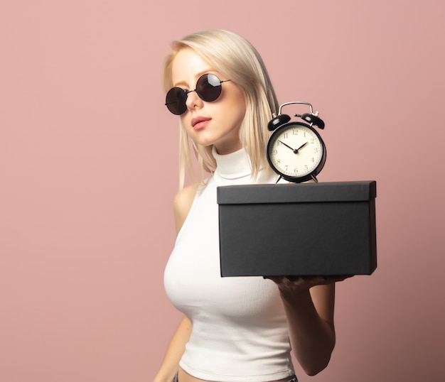 Style blonde in top and sunglasses with alarm clock and black gift box on pink