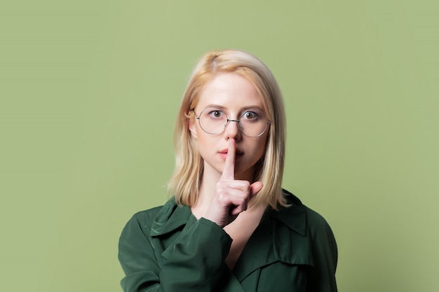 Style blonde woman in cloak and round glasses doing silence gesture