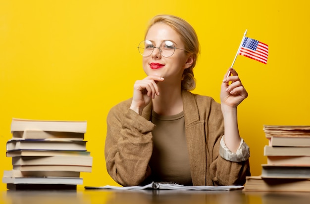 Photo style blonde woman sitting at table with books around and holds usa flag on yellow