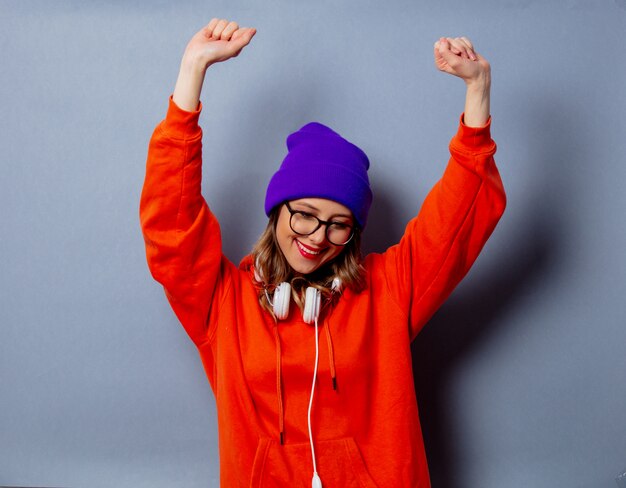 Style girl in orange hoodie and purple hat with headphones on grey wall