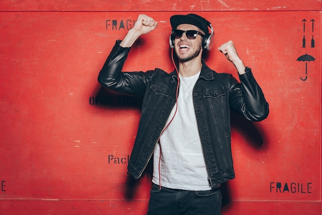 Style and music is his passion. Handsome young man in headphones gesturing and keeping mouth open while standing against red background