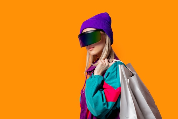 Style woman in 90s tracksuit and VR glasses with shopping bags on orange