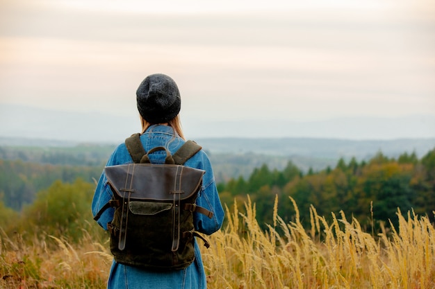 Style woman in denim jacket and hat with backpack in countryside with mountains