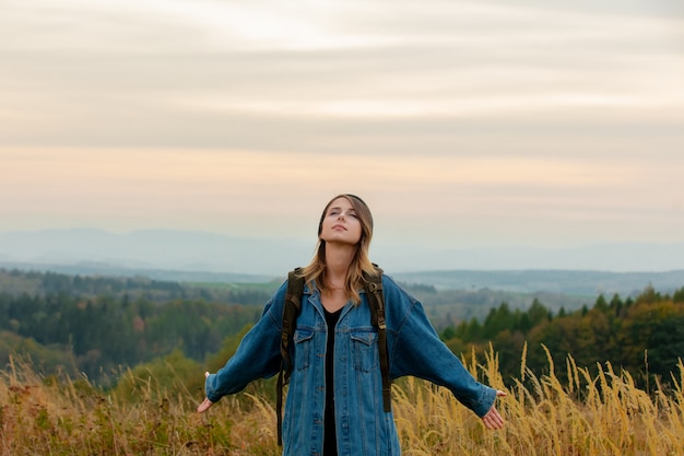 Photo style woman in denim jacket and hat with backpack in countryside with mountains