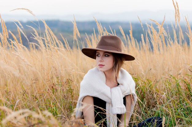 Style woman in sweater sits in yellow grass at countryside with mountains