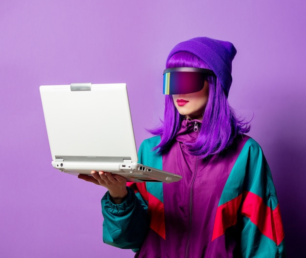 Style woman in VR glasses and 80s tracksuit with laptop on violet wall