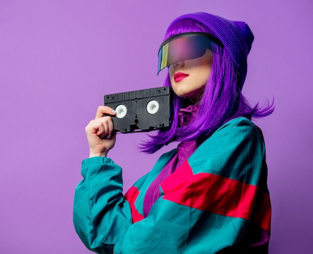 Style woman in VR glasses and 80s tracksuit with VHS tape on violet wall