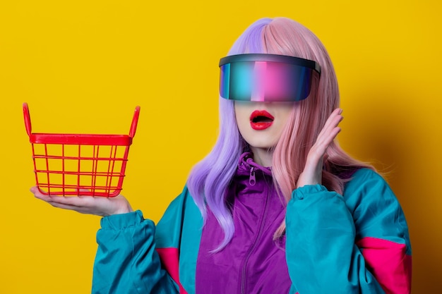 노란색 배경에 쇼핑 바구니가 있는 VR 안경과 90년대 스포츠 슈트를 입은 스타일 여성