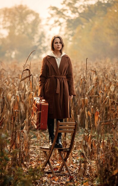 秋の季節にトウモロコシ畑の椅子にスーツケースとスタイルの女性滞在