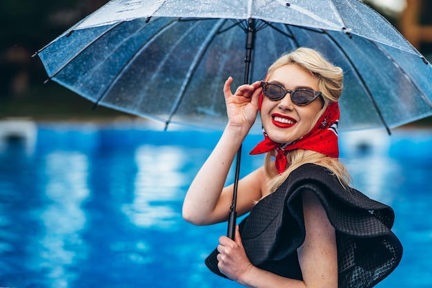 Styled blonde with sunglasses holding an umbrella in the swimming pool