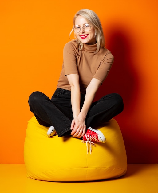 Styled hipster woman sitting in yellow bean chair on orange wall