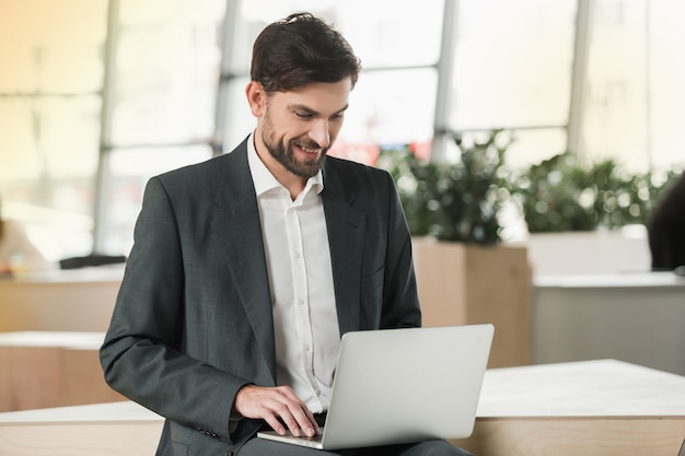 Stylish adult businessman while working day in office. Businessman working with laptop and smiling. Office interior with big window