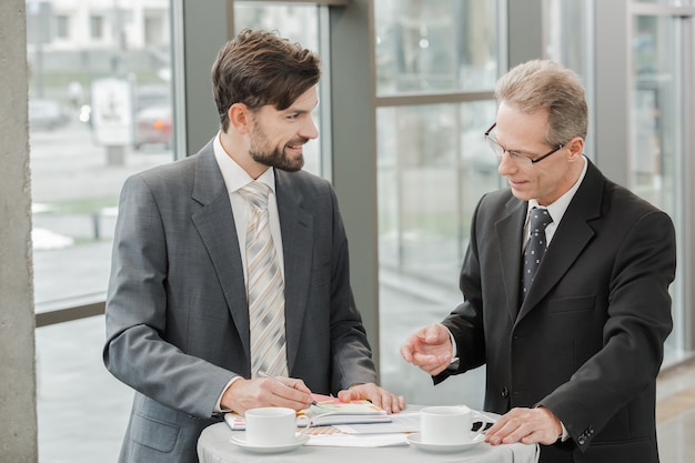 Stylish adult businessmen while working day in office. Businessmen talking and having coffee. Office interior with big window