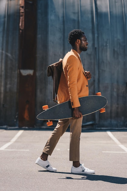 Stylish african american businessman holding longboard while walking on rooftop