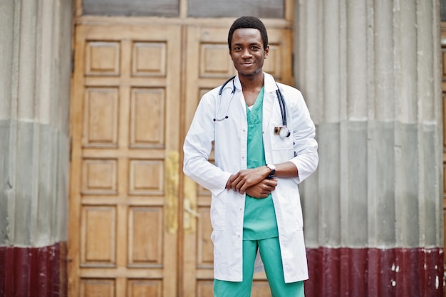 Stylish african american doctor with stethoscope and lab coat posed against door of hospital