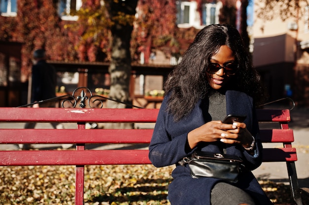 青いコートとサングラスでスタイリッシュなアフリカ系アメリカ人の女の子は、ベンチで晴れた秋の日にポーズをとって、彼女の携帯電話を使用しています。スマートフォンでアフリカモデルの女性。