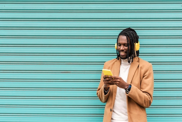 Stylish african american man in dreadlocks smiling and listening music with yellow headphones on blue background while sending a message with mobile phone
