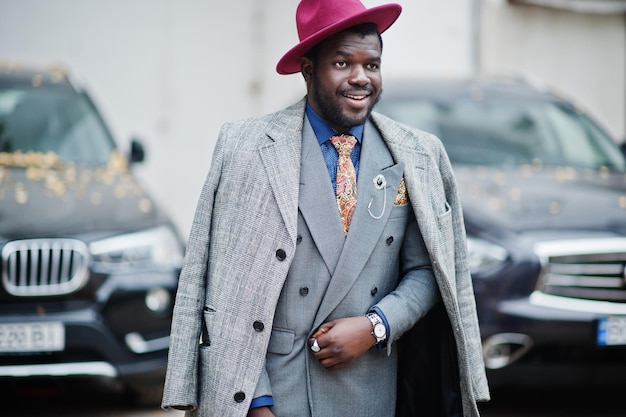 Stylish African American man model in gray coat jacket tie and red hat against two black business suv cars