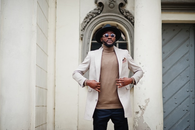 Stylish african american man wear beige jacket and  black hat with sunglasses pose against vintage window.