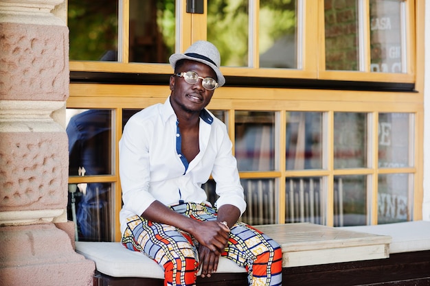 Stylish african american man in white shirt and colored pants with hat and glasses posed outdoor Black fashionable model boy