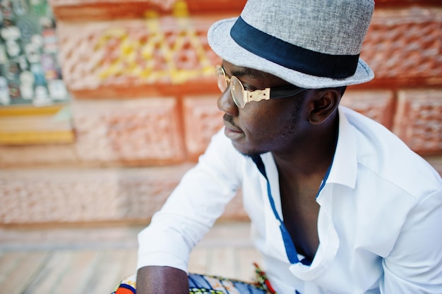 Foto elegante uomo afroamericano in camicia bianca e pantaloni colorati con cappello e occhiali in posa all'aperto ragazzo nero modello alla moda