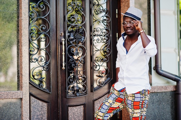 Stylish african american man in white shirt and colored pants with hat and glasses posed outdoor Black fashionable model boy