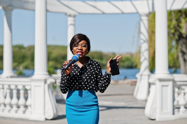 Stylish african american model girl TV presenter with microphone in glasses blue skirt and black blouse posed outdoor against white stone arch with mobile phone