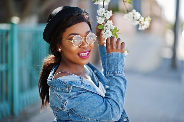 Elegante modello afroamericano in cappello occhiali, giacca di jeans e gonna nera poste all'aperto con alberi in fiore in primavera.