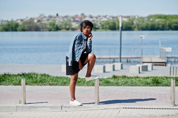 Stylish african american model in glasses hat, jeans jacket and black skirt posed outdoor.