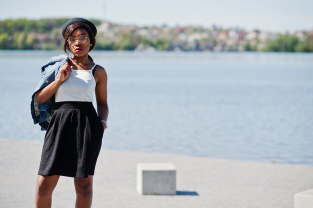 Stylish african american model in glasses hat, jeans jacket and black skirt posed outdoor.