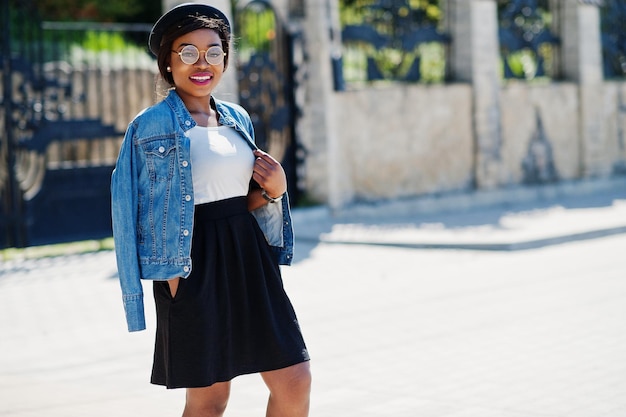 Стильная афроамериканская модель в очках, шляпе, джинсовой куртке и черной юбке позирует на улице