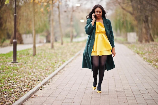 Stylish african american woman at green coat and yellow dress posed against autumn park and speaking on phone