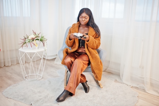 椅子に座っている部屋で提起され、古いビンテージ写真を保持しているオレンジ色のコートを着たスタイリッシュなアフリカ系アメリカ人女性