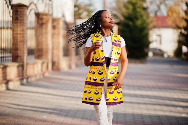 Stylish african american women in yellow jacket posed on street with hot drink in disposable paper cup