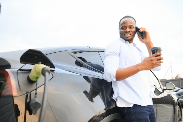 Стильный африканский мужчина с чашечкой кофе в руке вставляет выключатель в розетку зарядки электрического автомобиля