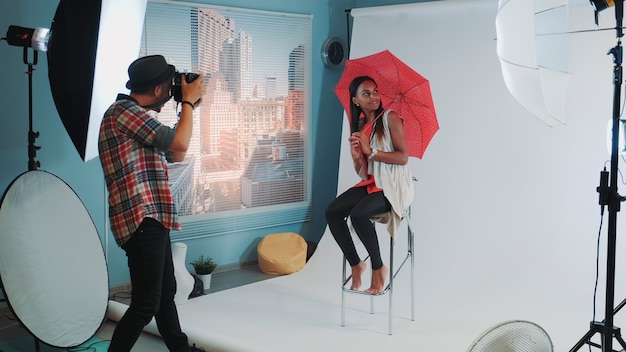 Stylish african model posing with red umbrella on bar high chair for fashion magazine photo shoot ph...