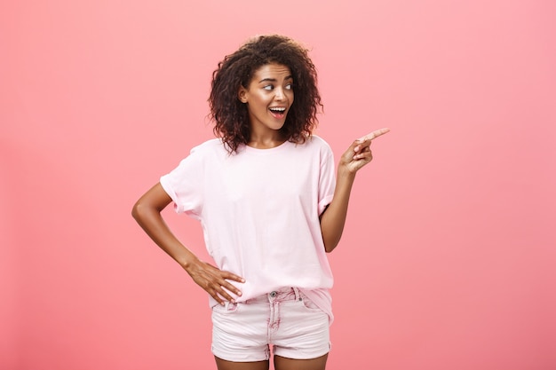 Elegante donna africana che punta e guarda a destra con sguardo curioso oltre il muro rosa