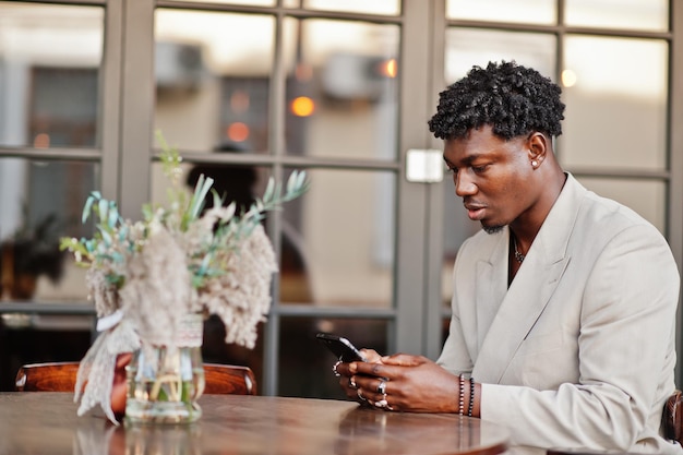 Stylish afro man in beige old school suit sitting on cafe with mobile phone Fashionable young African male in casual jacket on bare torso