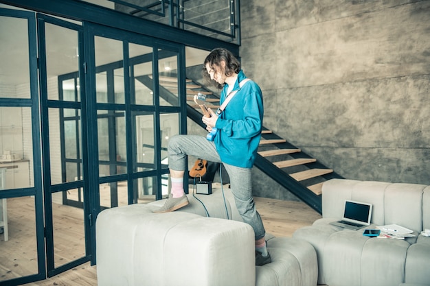 Stylish apartment. Pleasant inspired dark-haired guy standing on couch and actively playing while holding electronic guitar